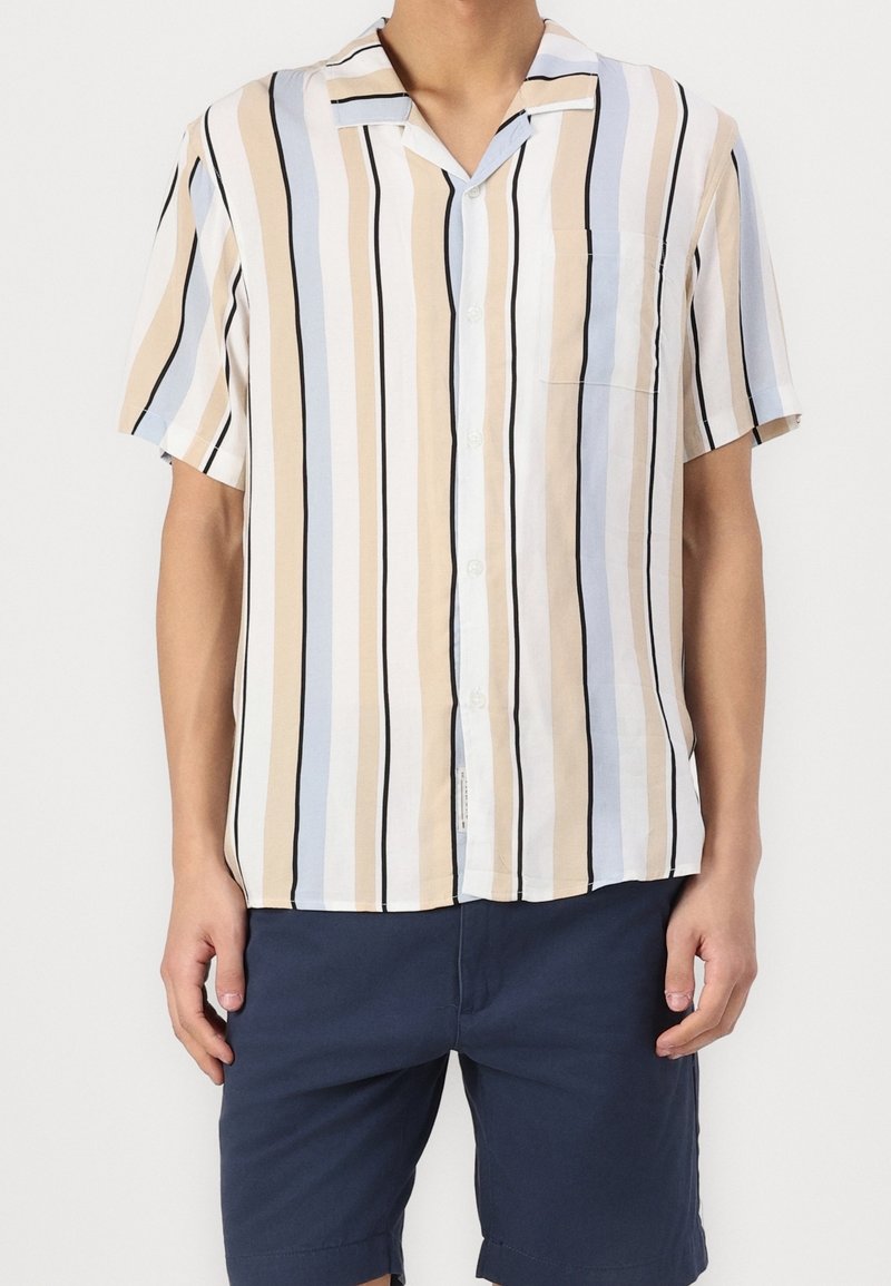 Homme portant une chemise à manches courtes à boutons avec des rayures verticales beige, bleu clair, blanc et noir, associée à un short bleu marine.