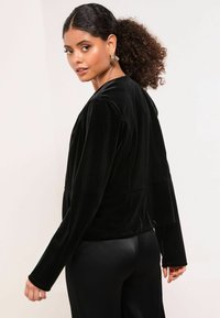 Černá sametová bunda s dlouhými rukávy, volným střihem a unikátními detaily na zadní straně. Hladká textura a minimalistický design.