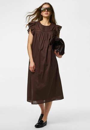 Femme portant une robe midi marron ample avec des manches à volants, des lunettes de soleil oversize, des mocassins noirs, tenant un sac à main en cuir marron foncé plié.