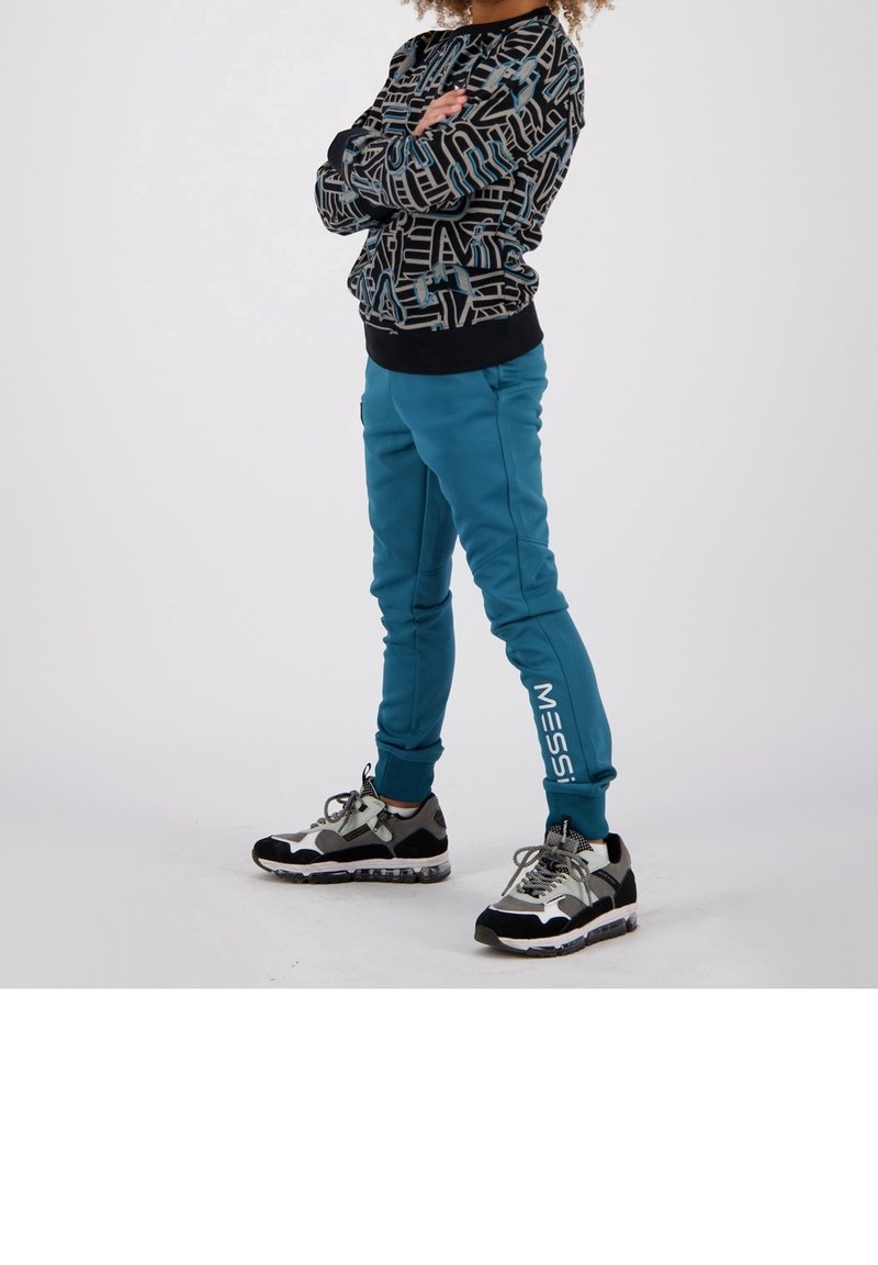 Enfant debout, bras croisés, portant un sweat à motifs noir et bleu, un pantalon de jogging bleu sarcelle avec le texte « MESSI », et des baskets noires et blanches.