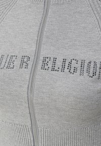 Gros plan sur un tissu tricoté gris clair avec une fermeture éclair verticale argentée et un texte clouté noir partiellement visible sur la poitrine.