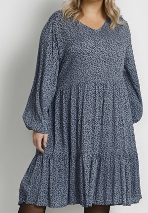 Femme portant une robe bleue ample jusqu'aux genoux avec de petits motifs floraux foncés et de longues manches bouffantes sur un fond uni.