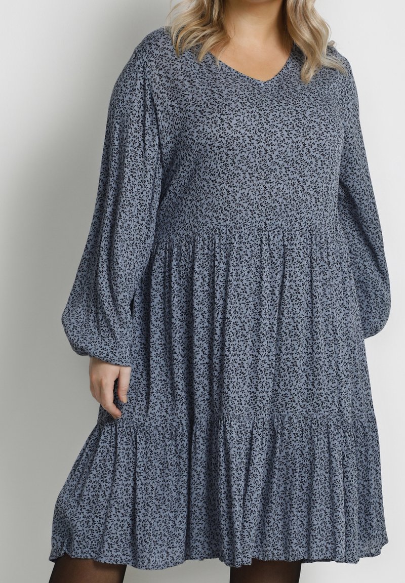 Femme portant une robe bleue ample jusqu'aux genoux avec de petits motifs floraux foncés et de longues manches bouffantes sur un fond uni.