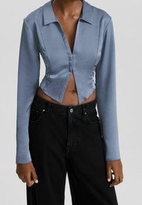 Blus i blå satin med lång ärm, krage och V-formad front, ihop med högmidjade svarta jeans i denim. Slät yta, kort skärning.