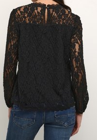 Svart spets topp med lång ärm som har intrikata blommönster, en vågig halsringning och ett nyckelhål bak med knappdetalj. Kombination med denim.