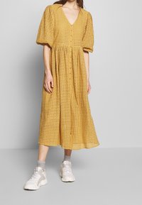 Senapsgul midiklänning med V-ringning, puffärmar och detaljer i ögonhålstygn. Matchas med vita sportskor och grå strumpor.