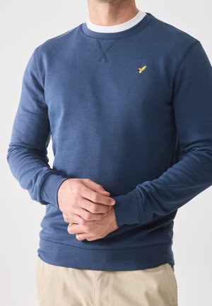 Mężczyzna ubrany w granatową bluzę z okrągłym dekoltem, z małym żółtym logo ptaka na piersi i beżowe spodnie, z rękami złożonymi na przodzie.
