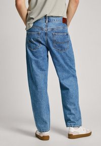 Jeans de mezclilla azul claro con un corte relajado, diseño de cinco bolsillos y parche en la parte trasera. Textura lisa y detalles de costura expuestos.