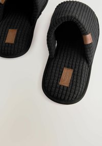 Chaussons en tissu noir avec une surface texturée, design ouvert sur le devant, et accents en cuir marron. Semelles en caoutchouc pour l'adhérence et la durabilité.
