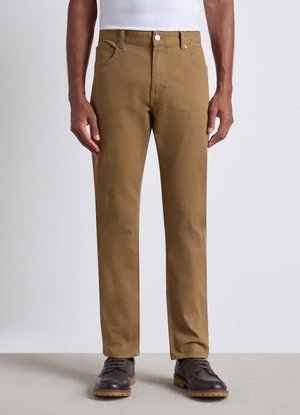 Mann trägt beige gerade geschnittene Hose, dunkelbraune Schnürschuhe und ein weißes Hemd vor einem einfarbigen hellen Hintergrund.