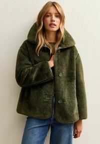 Kobieta w zielonej, oversize'owej kurtce z faux futra z dużym kołnierzem i niebieskich dżinsach, stojąca na tle gładkiej, jasnobeżowej ściany.