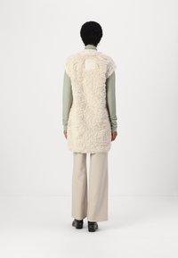 Un lungo gilet beige chiaro realizzato in un materiale soffice e testurizzato, con collo rotondo e senza chiusure, indossato sopra una maglietta verde e pantaloni beige chiaro.