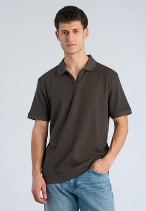 Junger Mann mit kurzen dunklen Haaren, der ein dunkelbraunes Poloshirt mit kurzen Ärmeln und hellblaue Jeans trägt, steht mit einer Hand in der Tasche.