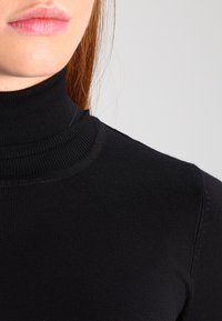 Czarny top z wysokim, przypominającym golf kołnierzem, o prążkowanej fakturze i dopasowanym kroju, wykonany z gładkiej tkaniny z delikatnymi szwami w pobliżu kołnierza.