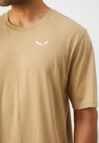 T-shirt in cotone beige con maniche corte, dotato di un logo bianco sulla parte superiore sinistra del petto. Il tessuto appare morbido e liscio con una vestibilità attillata.