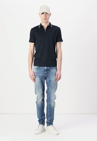 Tricou polo bleumarin cu accente albe, asortat cu blugi deschis la culoare, deteriorați, și adidași albi. Figură masculină purtând o șapcă bej.
