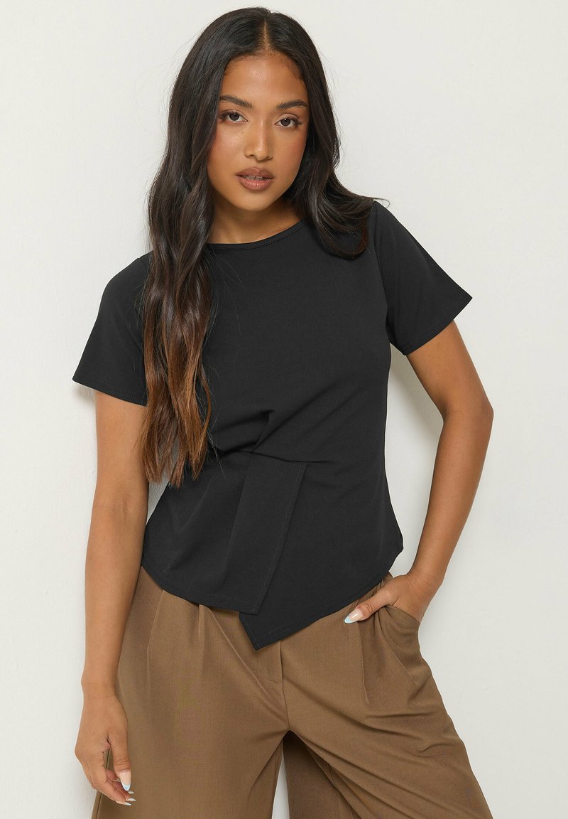 Femme aux longs cheveux foncés portant un haut noir à manches courtes avec un ourlet asymétrique et un pantalon marron taille haute, posant avec une main dans la poche.