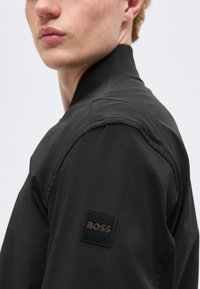 Νεαρό άτομο που φοράει μαύρο μπουφάν με τετράγωνο έμβλημα στην επάνω πλευρά του βραχίονα, όπου αναγράφεται "BOSS", κοιτάζοντας πλάγια, με απλό φόντο.