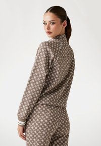 Braun gemusterter Trainingsanzug mit weißen Details, der einen hohen Kragen und gestreifte Bündchen aufweist. Der Stoff hat eine glatte Textur und ein wiederkehrendes Design.