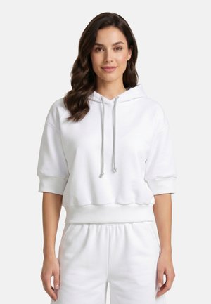 Vrouw draagt een witte cropped hoodie met koordjes en bijpassende witte joggingbroek, staand tegen een effen lichte achtergrond.
