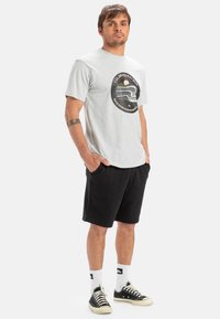 Camiseta gris de manga corta con un diseño gráfico circular, combinada con pantalones cortos negros. El modelo lleva zapatillas negras y calcetines blancos con logotipos.