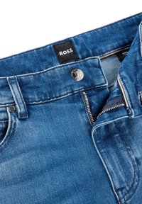 Närbild av blå denimjeans med en delvis uppdragen dragkedja, en metallknapp och en svart tygetikett med texten "BOSS" inuti midjebandet.