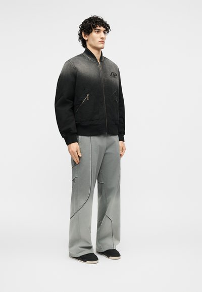 Νεαρός άνδρας με σγουρά μαλλιά, φοράει μαύρο μπομπέρ μπουφάν ombre και χαλαρό γκρι παντελόνι με μαύρα παπούτσια, στέκεται μπροστά σε ένα απλό φόντο.