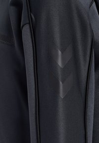 Svart sportjacka med en slät textur, försedd med en logotyp och tre svarta vinklar på ärmen.