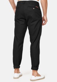 Pantalon en lin noir à coupe fuselée, ourlets retroussés, deux poches arrière et une étiquette en cuir avec logo sur la ceinture. Porté avec des chaussures blanches.