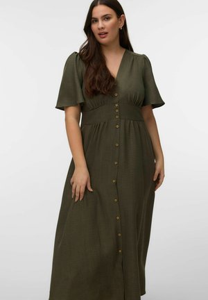 Ženska z dolgimi temnimi lasmi, oblečena v olivno zeleno midi obleko s kratkimi rokavi in gumbi, pred enostavnim ozadjem.