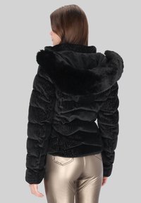 Czarna pikowana kurtka z kapturem wyściełanym futrem, teksturowaną tkaniną i poziomym przeszyciem, noszona z metalicznymi złotymi spodniami.