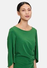 Grünes Kleid mit weichem, fließendem Stoff, weitem Ausschnitt und drapierten Ärmeln, ausgestattet mit gerafften Details an der Taille.