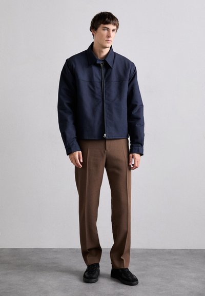 Navy blue jacket with a zip front, collared design, and two side pockets, paired with brown trousers and black sneakers. Smooth texture.