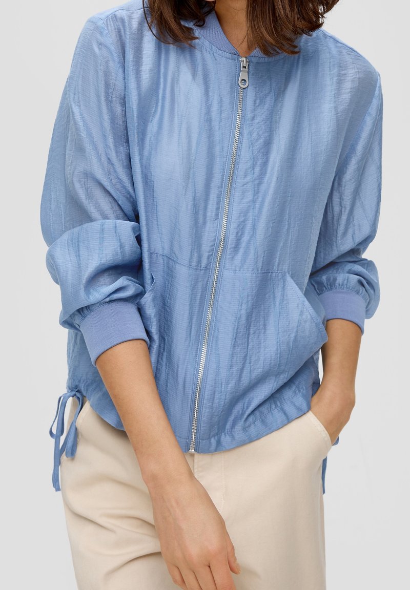 Femme portant une veste bleu clair à fermeture éclair avec des poignets élastiques et un pantalon beige, une main dans la poche, sur fond uni.