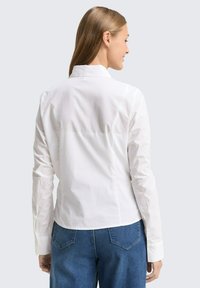 Witte langslavige shirt met een kraag, gemaakt van lichte stof. Heeft achterste naden en een licht cropped snit, gecombineerd met blauwe denim jeans.