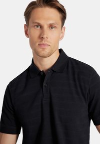 Młody mężczyzna z jasnobrązowymi włosami noszący czarną, strukturalną koszulę polo z krótkim rękawem na białym tle.