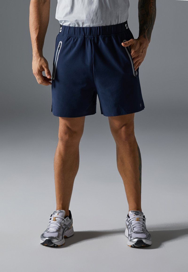 Marineblå shorts med elastisk talje, sidelommer med lynlås og en glat overflade. Kombineret med grå og sorte træningssko.