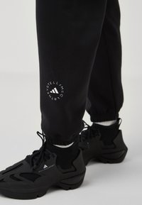 Dolní končetiny v černých teplácích Stella McCartney a černých teniskách Adidas Stella McCartney s černými tkaničkami na šedé podlaze.