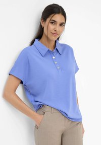 Polo di colore azzurro chiaro con colletto e maniche corte, dotato di una pattina con quattro bottoni argentati. Tessuto liscio e strutturato.