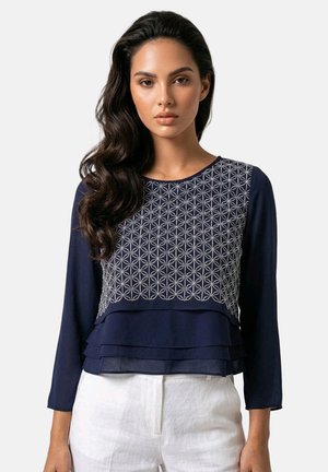Mujer con cabello oscuro largo y ondulado que lleva una blusa azul marino de manga larga con bordado geométrico blanco y pantalones blancos, mirando al frente.