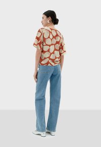 Kurzärmlige, rot-beige gemusterte Bluse, kombiniert mit hellblauen Jeans. Der Stoff sieht weich aus, hat eine lockere Passform und einen Rundhalsausschnitt.