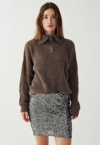 Brūns ribots džemperis ar augstu apkakli un rāvējslēdzēja detalizāciju, uzvilkts kopā ar pieguļošu melnu spīdīgu mini svārku, kam ir krokotu akcenti.