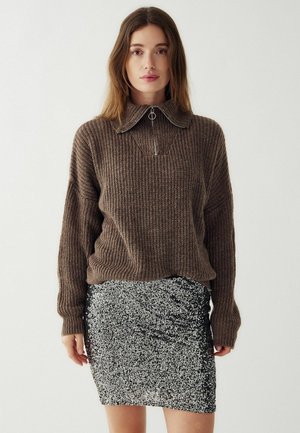 Barna bordás pulóver magas gallérral és cipzáras részletekkel, amit egy testhez simuló fekete flitteres mini szoknyával párosítottak, ruched díszítésekkel.