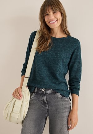 Dunkeltürkis gestrickter Pullover mit langen Ärmeln und subtiler Textur, kombiniert mit grauen Jeans und einer hellcremefarbenen, lässigen Handtasche.