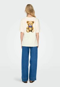 T-shirt couleur crème avec un graphique d'un ours en peluche marron portant un nœud papillon bleu au dos. Coupe décontractée avec des manches courtes.