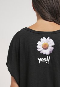 Černé tričko s pohodlným střihem, s bílou grafikou sedmikrásky a textem "ano!!" vytištěným pod květinou na zadní straně.