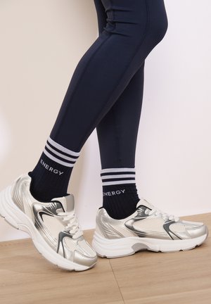 Sneakers atletici argento con dettagli in rete, suole bianche e accenti neri, abbinati a calze sportive a righe blu scuro e leggings.