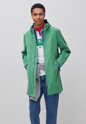 Junger Mann trägt grüne Kapuzenjacke, rotes Bandana, weißes grafisches T-Shirt, Jeansjacke, blaue Jeans und grauen Sweatshirt um die Taille gebunden.