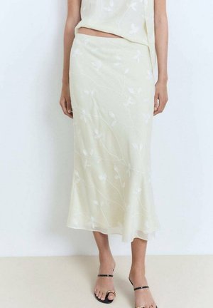 Woman wearing a cream midi skirt with subtle floral embroidery and a matching top, paired with black strappy sandals.