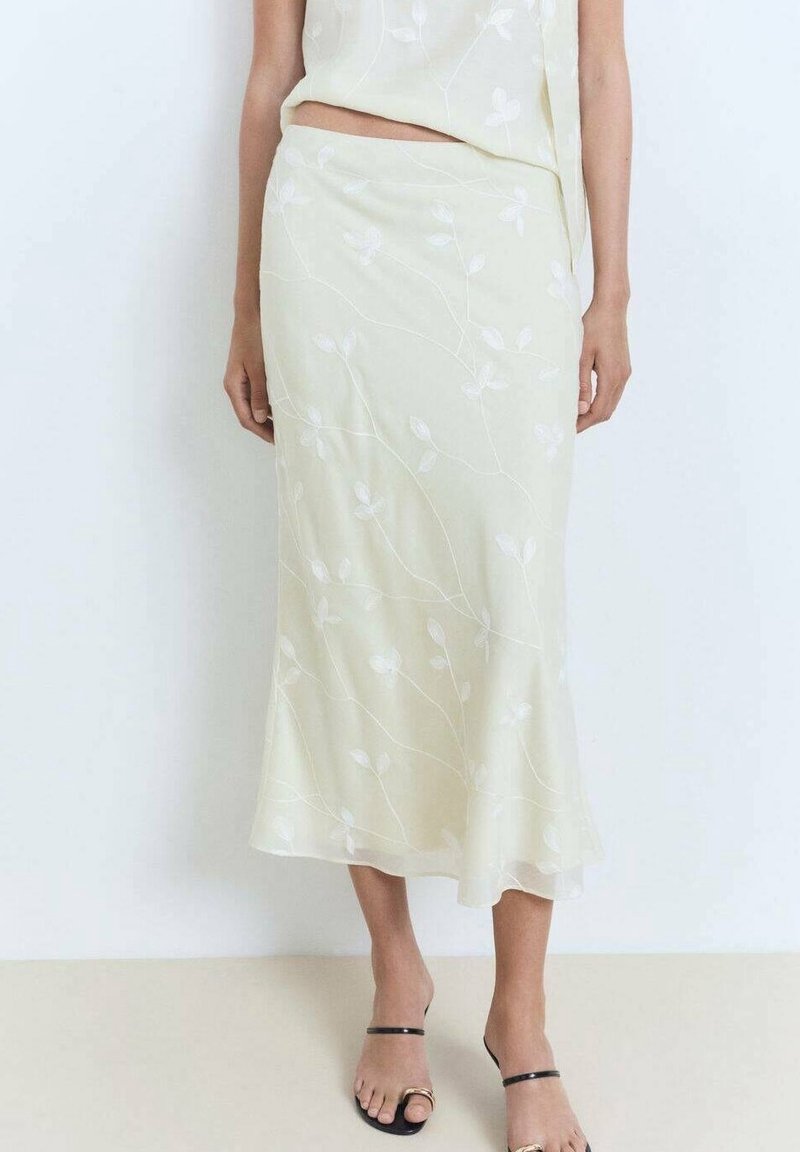Femme portant une jupe midi crème avec une broderie florale subtile et un haut assorti, associée à des sandales noires à brides.